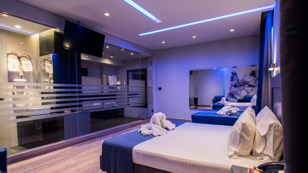 A modern hotel room with a large bed, a TV, a mirror, and a bathroom with a glass partition. - Pines Hotel (Kifisia)