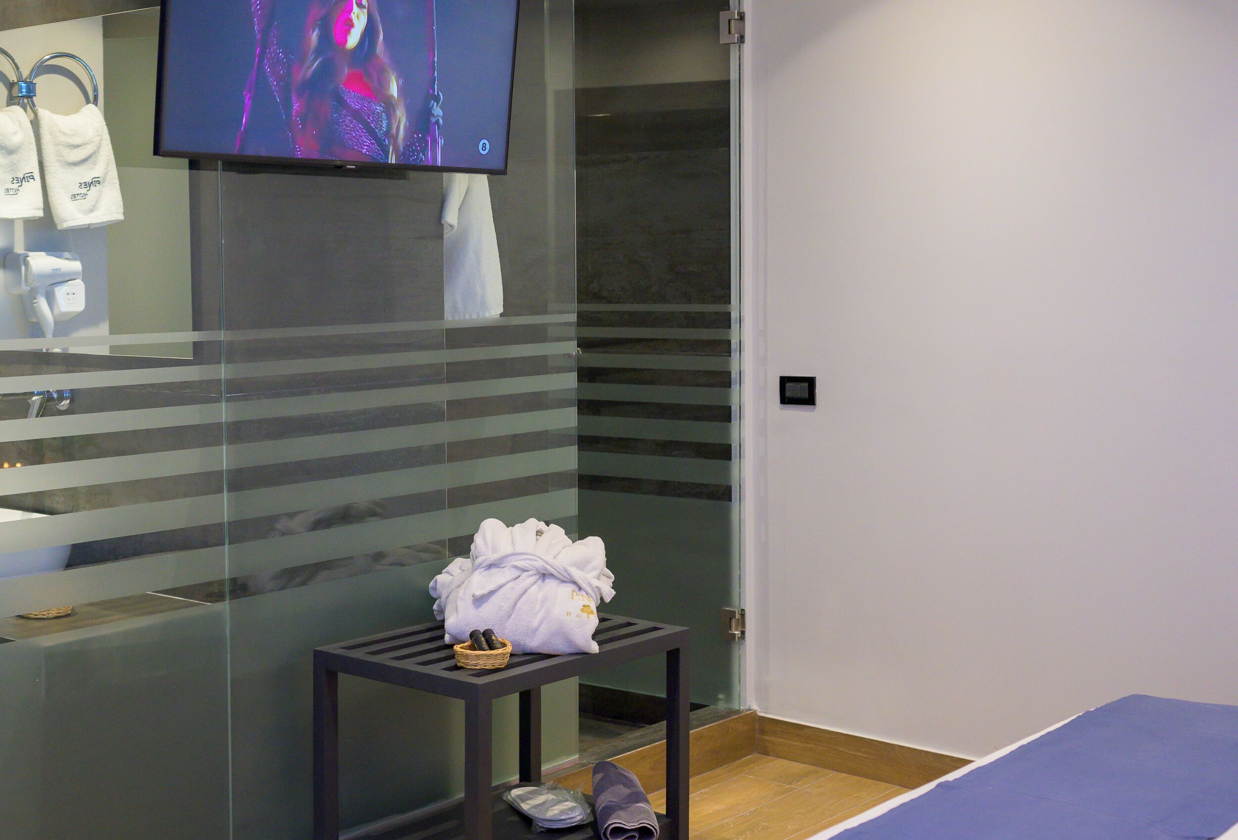 A modern bathroom with a glass shower enclosure, a towel rack with towels, a television mounted above the shower, and a small table with a towel and a basket.