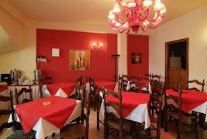 A dining area with red walls, multiple tables with white tablecloths, wooden chairs, and a chandelier. - Varonos (Delphi)