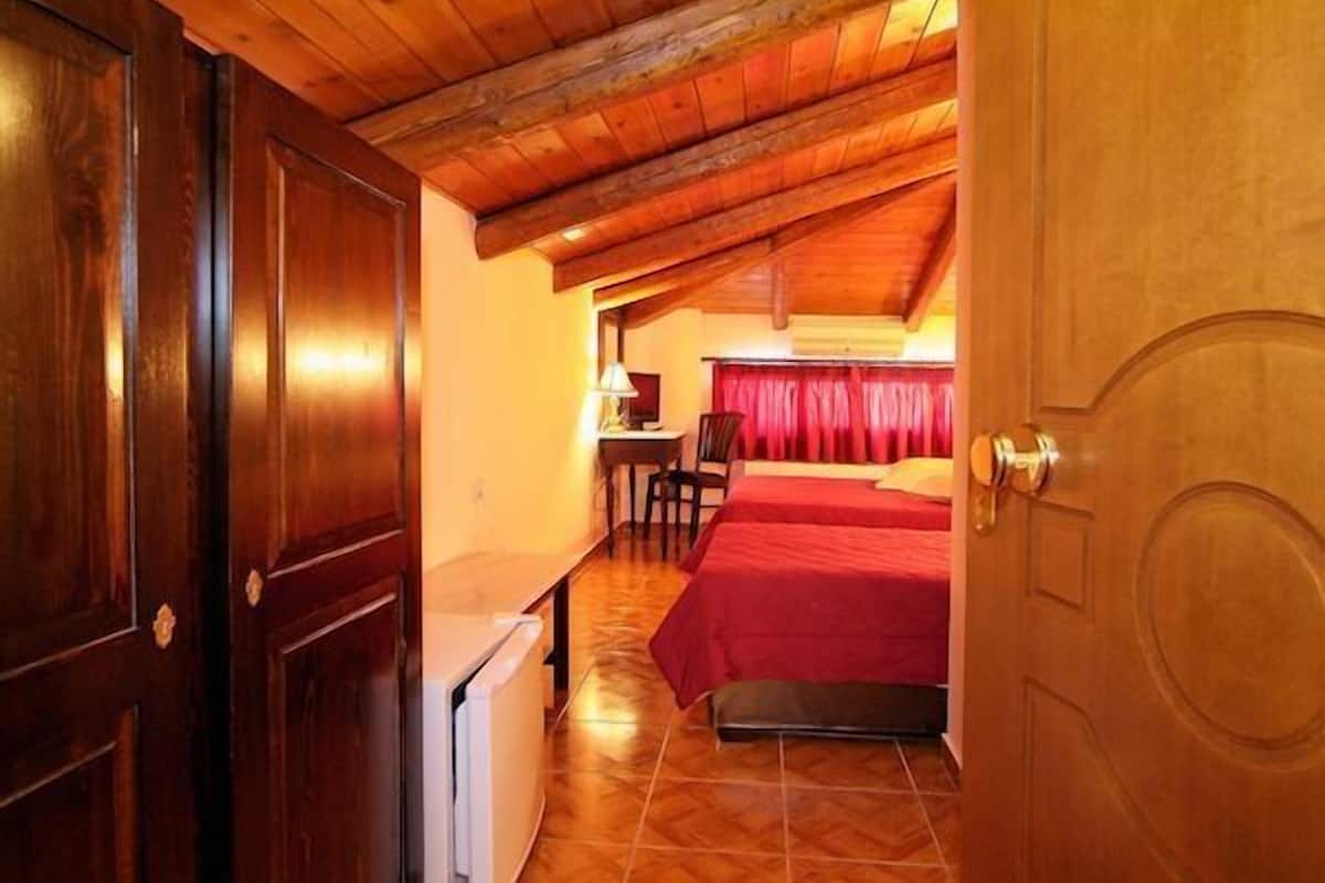 A room with a wooden ceiling, a bed with red bedding, a small table with a lamp, and a chair.