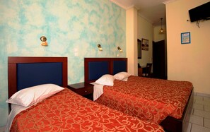 A hotel room with two beds, each with a blue headboard and red patterned bedspread. There are wall sconces with light bulbs, a flat-screen TV mounted on the wall, and a framed picture above the door. - Pergamos (Athens)