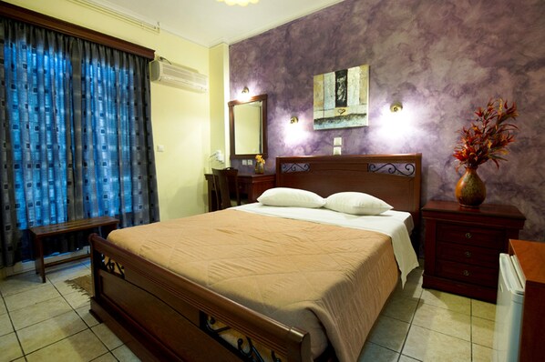 A hotel room with a large bed, a nightstand, a dresser, a mirror, and a window with curtains. - Pergamos (Athens)