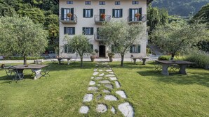 A multi-story building with a stone pathway leading to the entrance, surrounded by lush greenery and trees. - Crotto Regina (Gravedona)