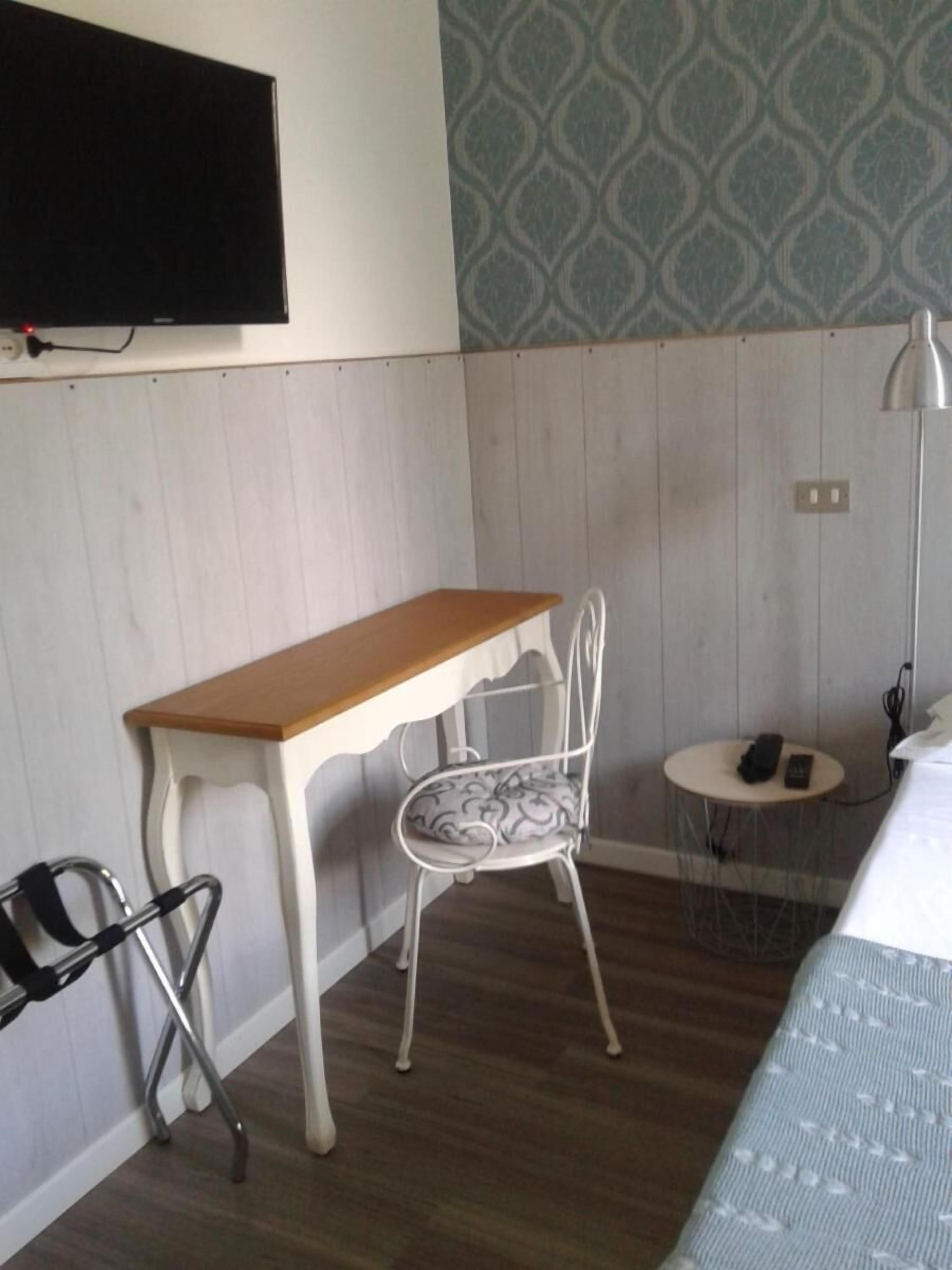 A hotel room with a wooden desk, a white metal chair, a small round table with a phone, and a bed with a patterned bedspread.