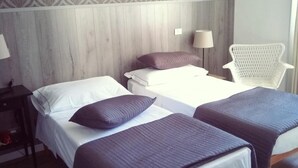 A hotel room with two beds, a wooden headboard, patterned wallpaper, a white chair, and a nightstand with a lamp.