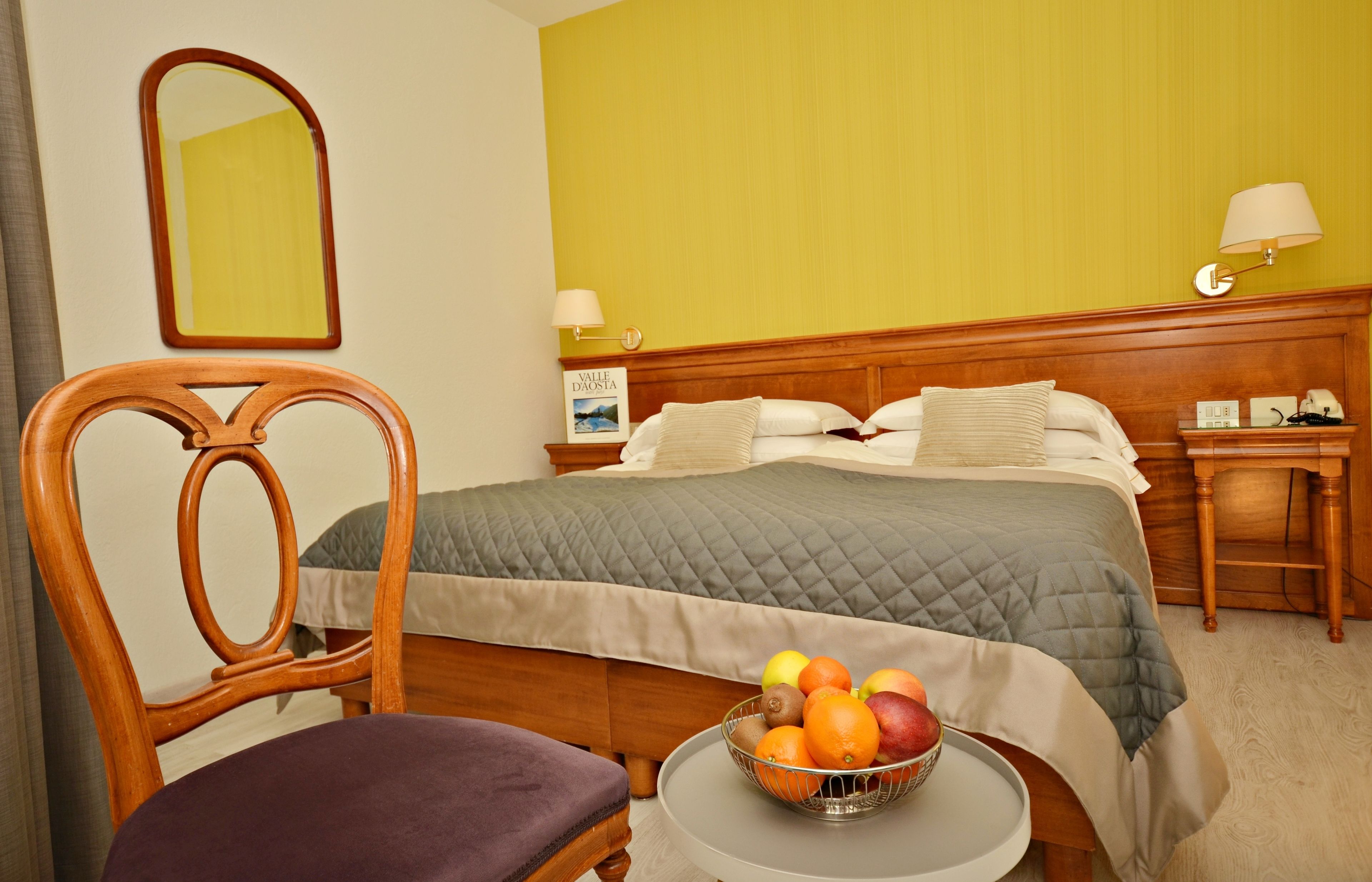 Ein Hotelzimmer mit Bett, Sessel, Nachttisch, Lampe und Obstschale auf einem kleinen Tisch.