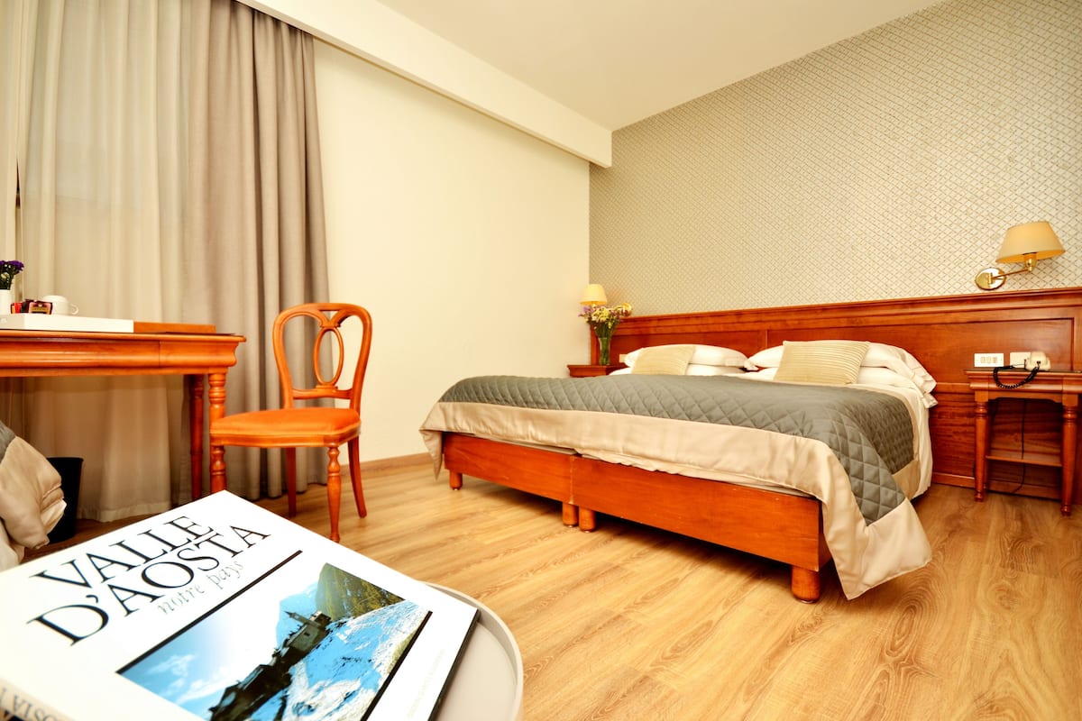 Habitación de hotel con cama de madera, un escritorio y una silla. Hay un libro sobre el escritorio y una lámpara en la mesita de noche.
