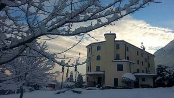 Un edificio de varios pisos con techo plano y una chimenea, rodeado de árboles cubiertos de nieve y una montaña al fondo.