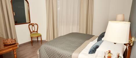 A hotel room with a bed, a chair, a nightstand, a lamp, a mirror, and a window with curtains.