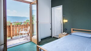 A bedroom with a bed, a nightstand, a telephone, a radiator, a wooden chair, and a sliding glass door leading to a balcony with a view of mountains.
