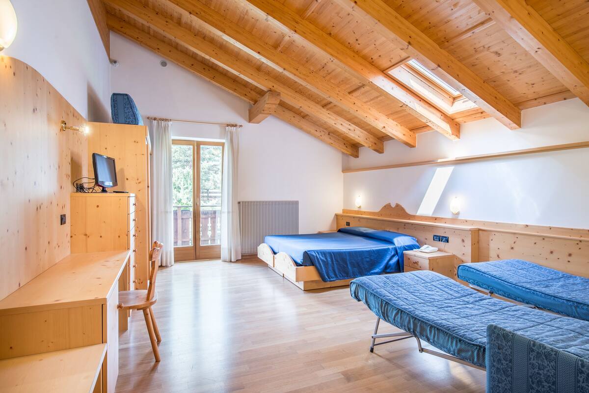 Une pièce avec un plafond en bois, un lit recouvert d’une couverture bleue, un bureau avec une chaise et une fenêtre avec des rideaux.