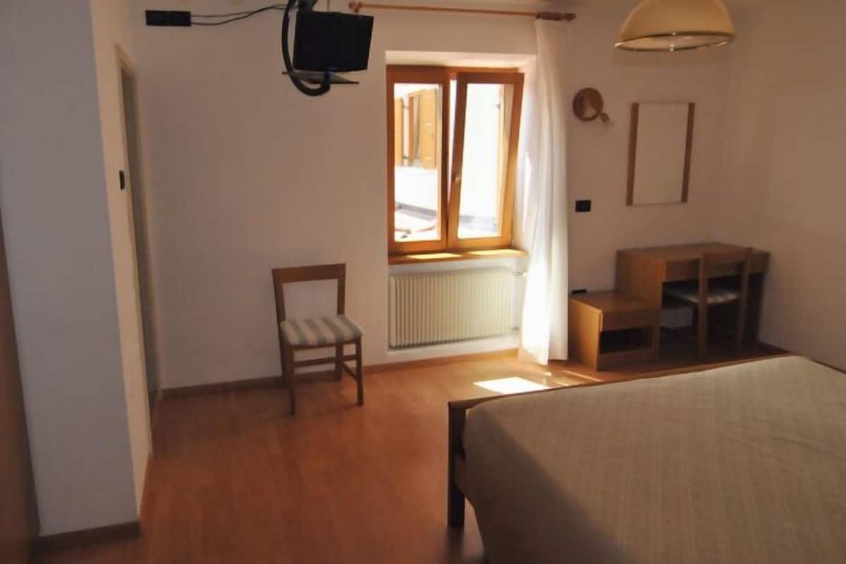Une pièce comprenant un lit, une chaise, une petite table, une fenêtre avec des rideaux, un radiateur et un téléviseur fixé au mur.