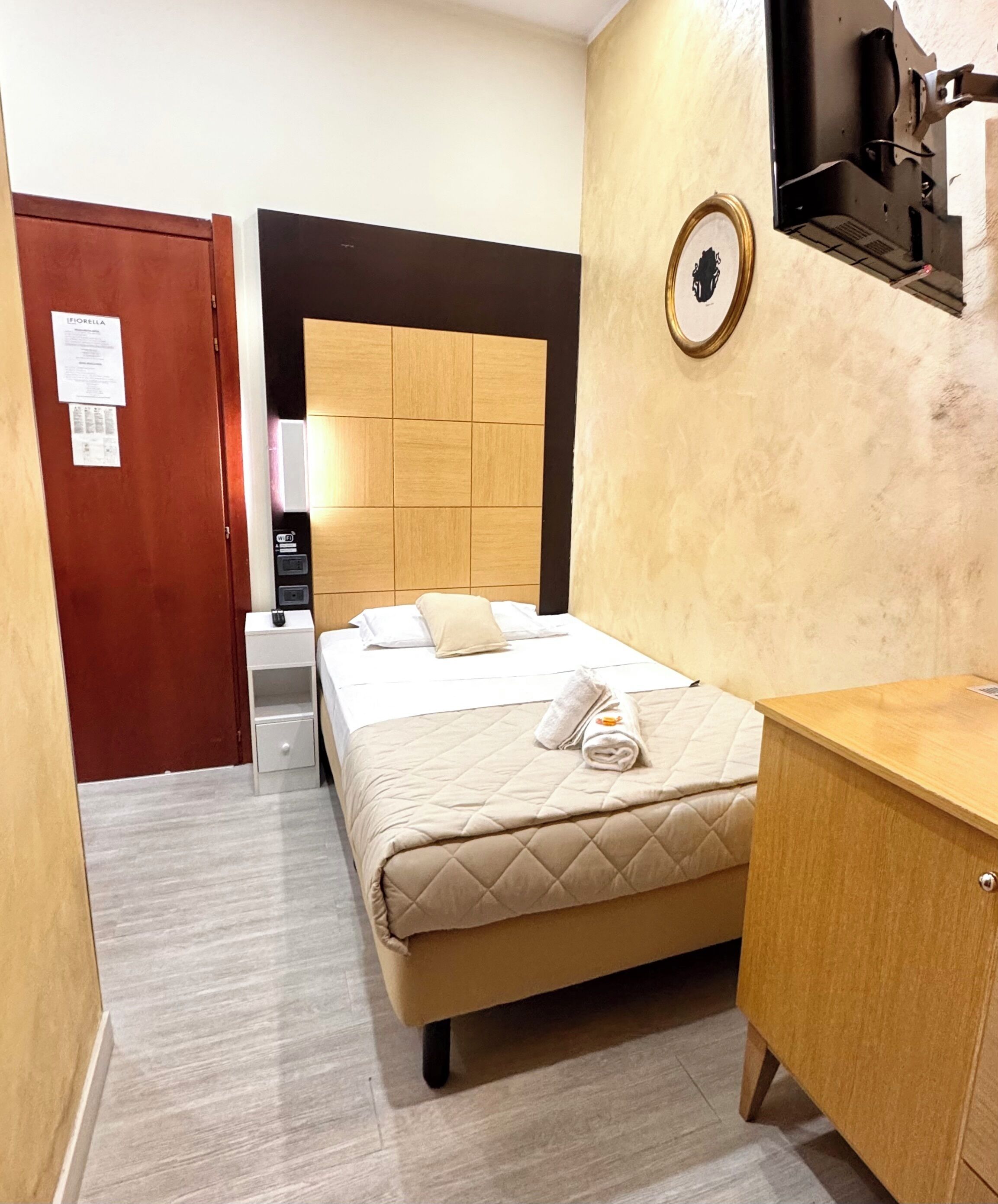 Ein Hotelzimmer mit einem Bett, einem Holz-Kopfteil, einem Nachttisch, einem Kleiderschrank und einem an der Wand montierten Fernseher.