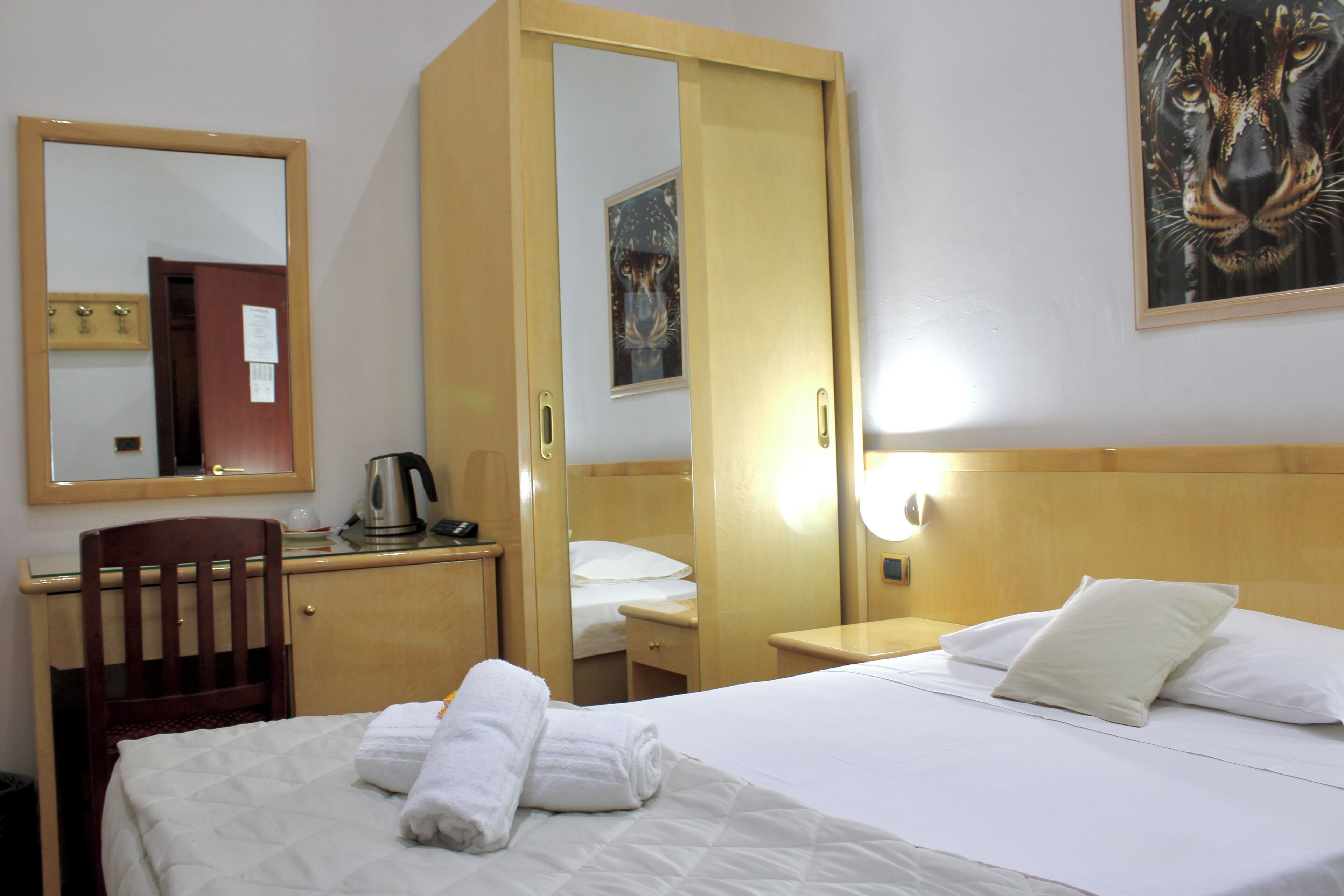 Ein Hotelzimmer mit Bett, Schreibtisch mit Stuhl, Spiegel, Gemälde und Nachttisch mit Lampe.