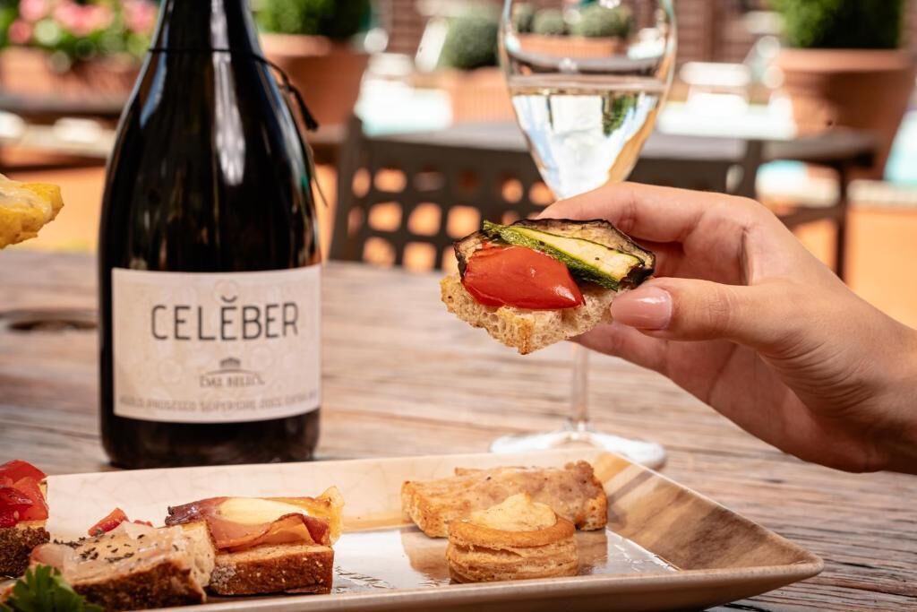 Een hand houdt eten vast, met een fles waarop 'CELEBER' staat en een glas witte wijn op een tafel.