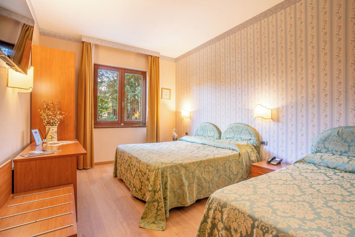 A hotel room with two beds, a wooden desk, a window with curtains, and floral wallpaper.