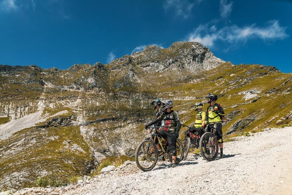 Ciclistas de montanha em uma trilha pedregosa, com montanhas ao fundo.