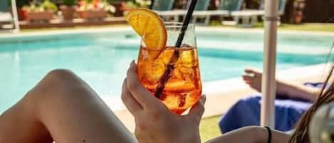 A person relaxing by a pool with a drink, surrounded by greenery and a mountain view.