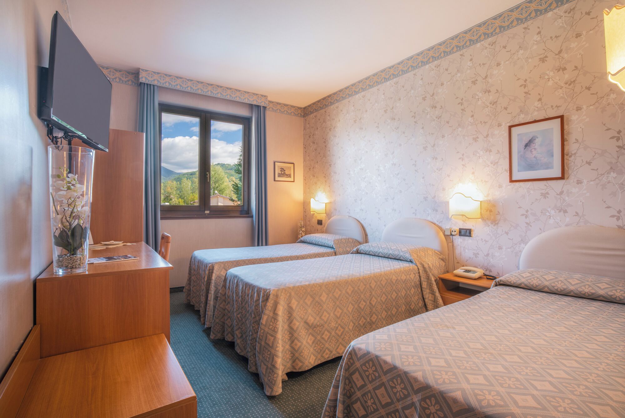 Ein Hotelzimmer mit zwei Betten, einem Schreibtisch, einem Fernseher und einem Fenster mit Blick auf Grünflächen.