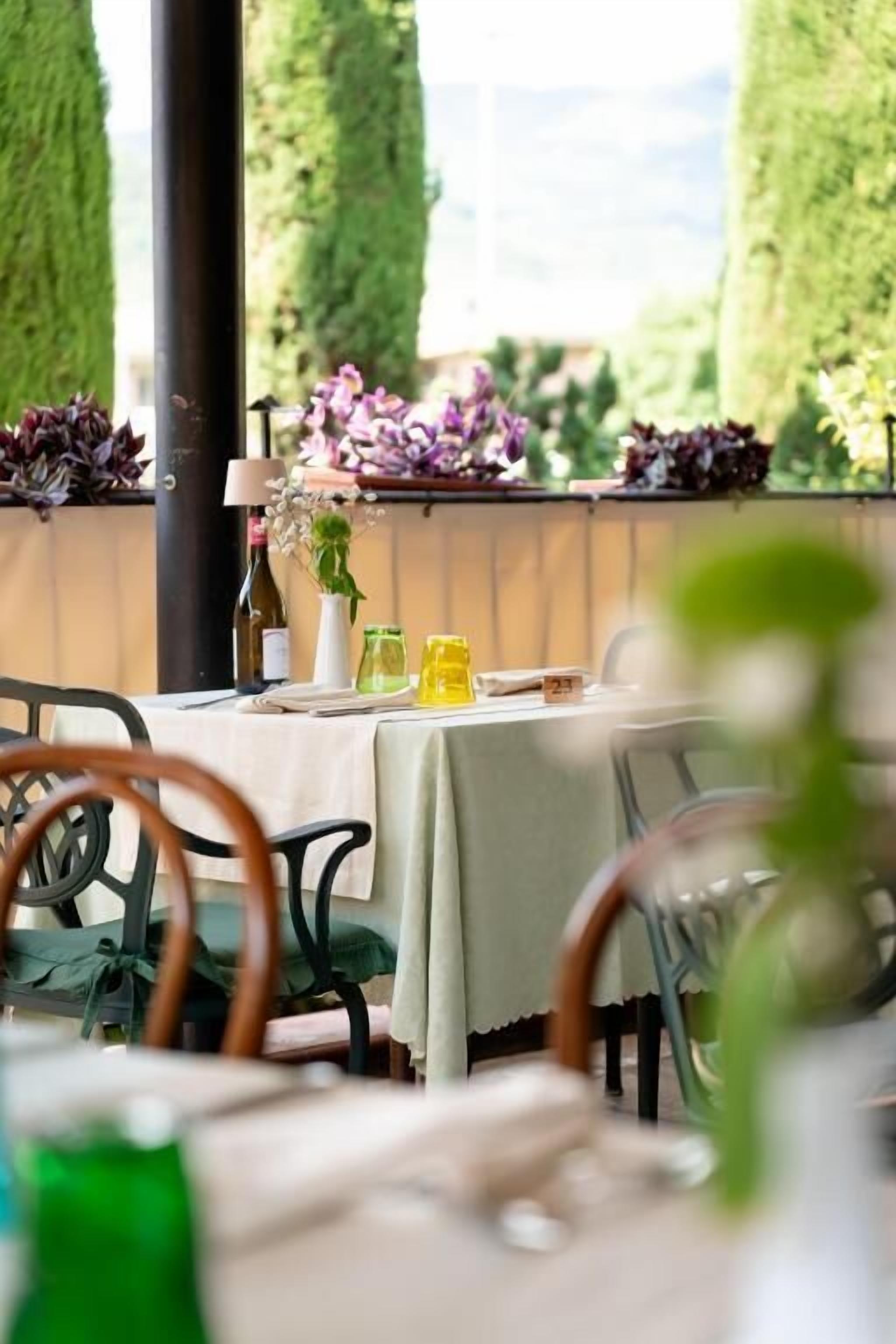 An outdoor dining area with tables set for two, featuring a tablecloth, a wine bottle, and a vase with flowers.
