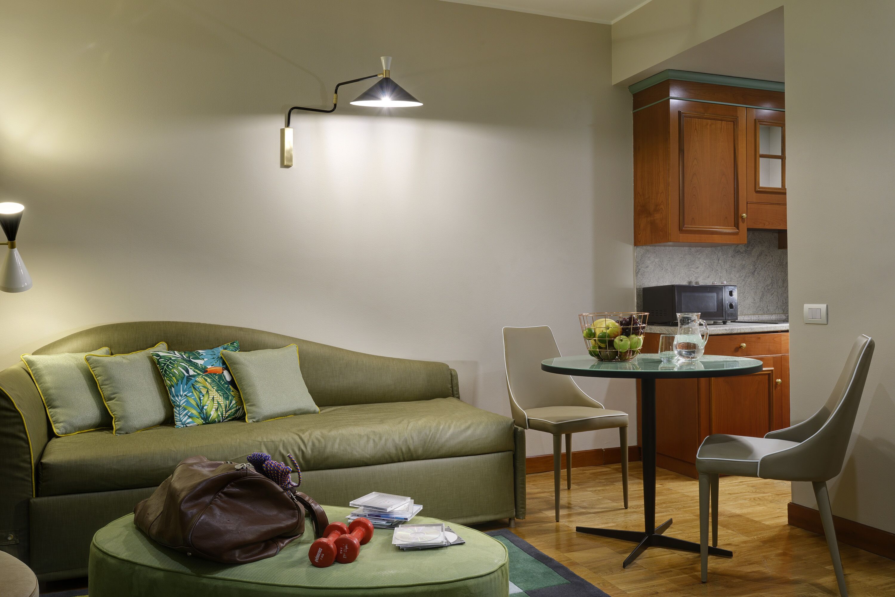 A compact living space with a sofa, ottoman, and a small dining table with chairs. A kitchen area is visible in the background.