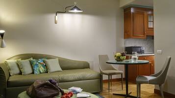 A compact living space with a sofa, ottoman, and a small dining table with chairs. A kitchen area is visible in the background.