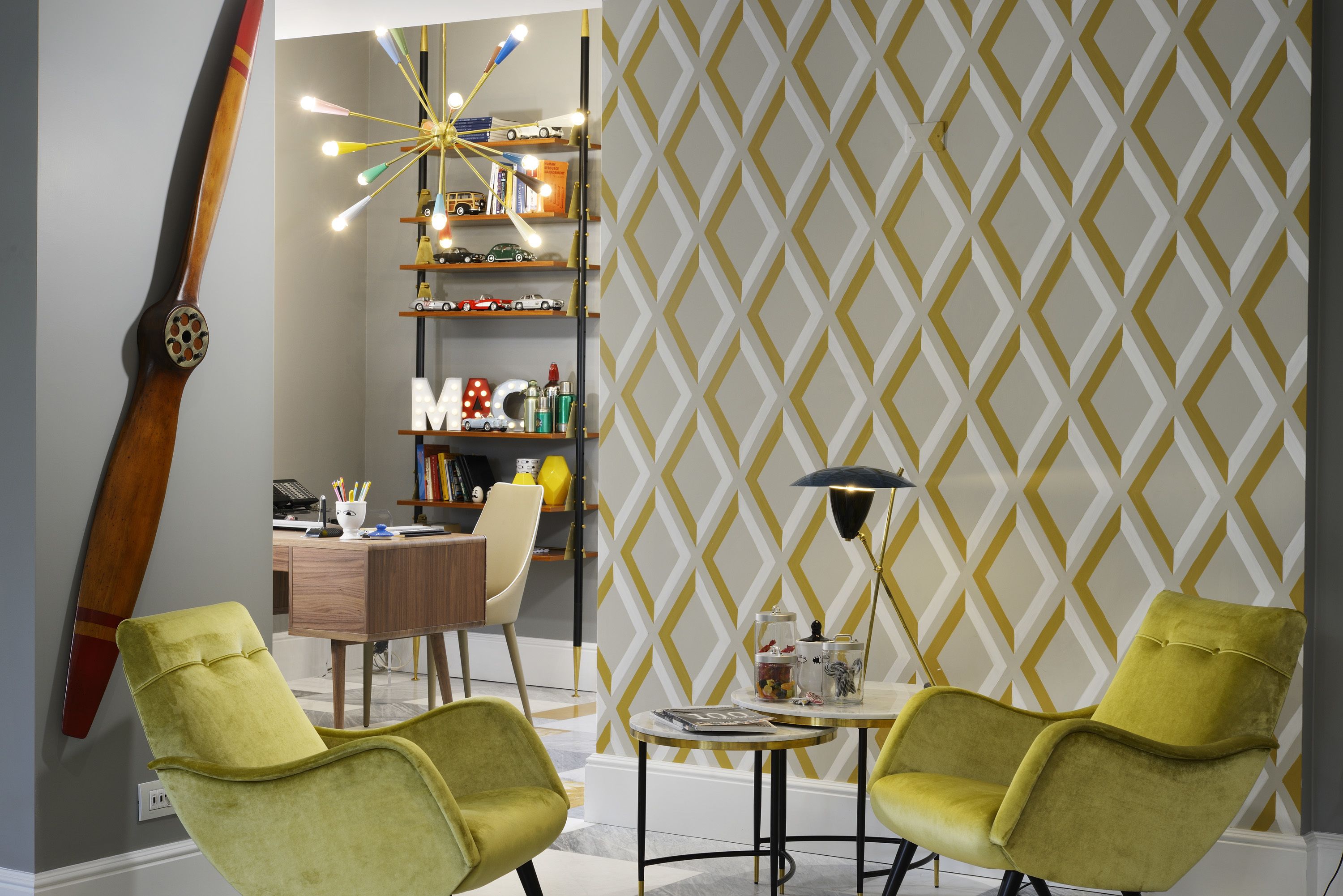 Una habitación de diseño moderno, con un escritorio, dos sillas verdes, una estantería llena de juguetes y un papel pintado estampado.