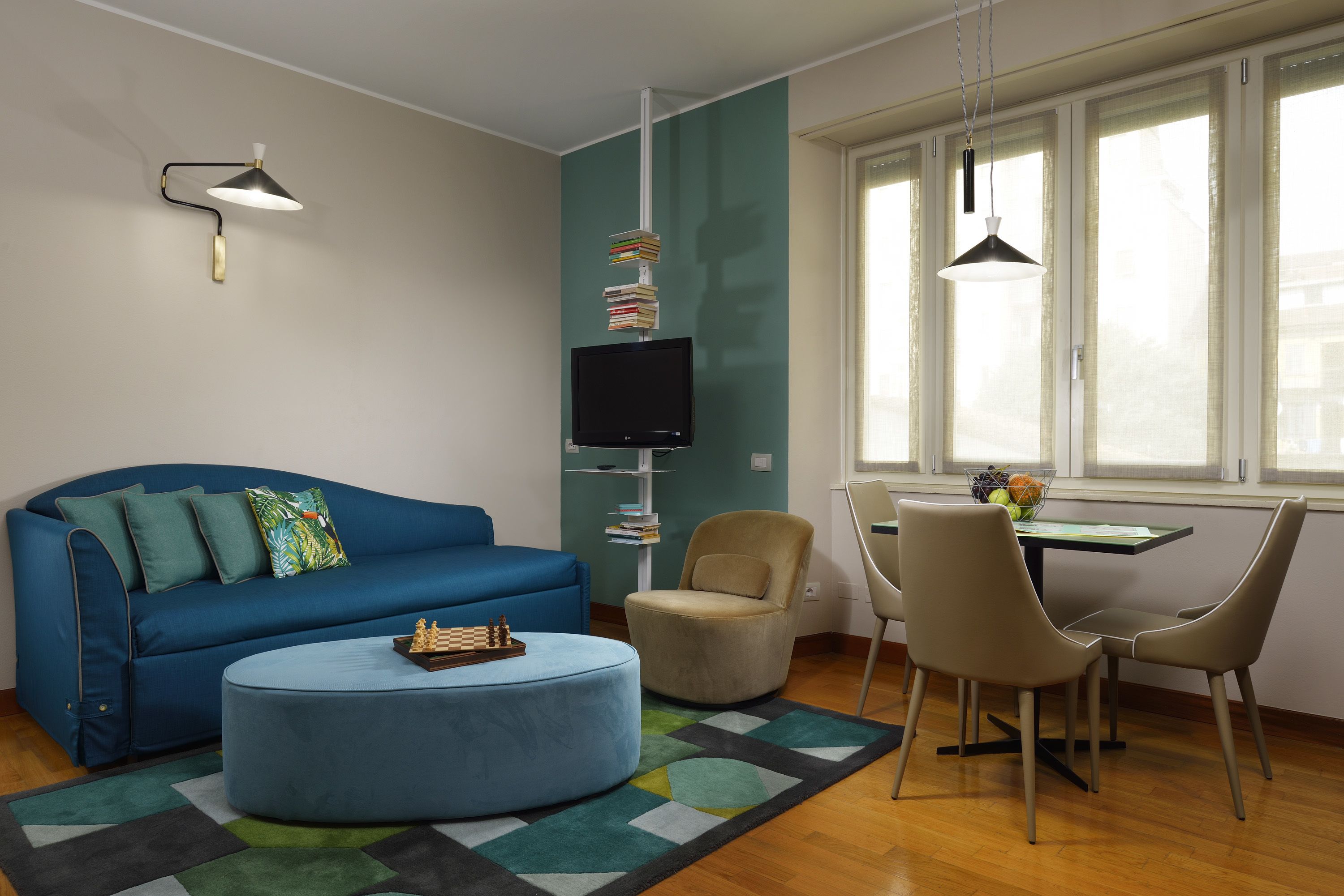 A modern living room with a blue sofa, a round ottoman, a dining table with chairs, a TV on a stand, and a bookshelf.