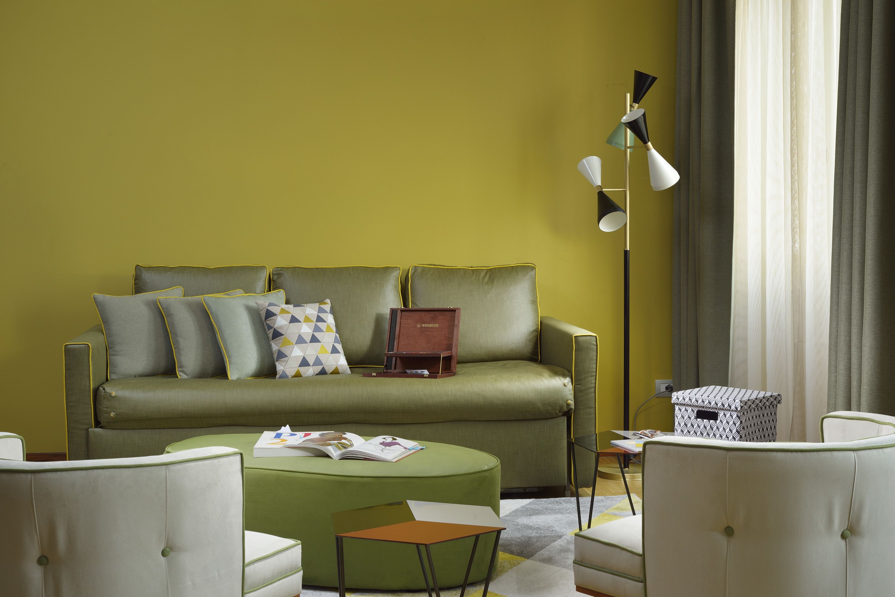 A living room with a green sofa, a small round table, and two armchairs. There is a floor lamp and a window with curtains.