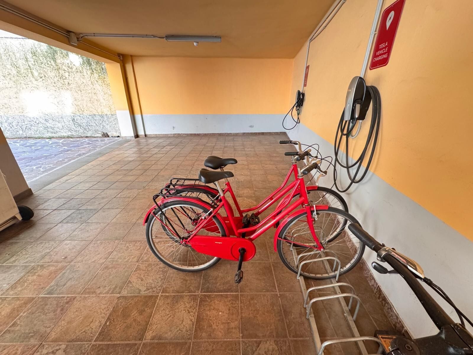 Ein rotes Fahrrad mit Korb und Sitz, abgestellt in einem Innenraum mit Fliesenboden und einer an der Wand befestigten Ladestation.