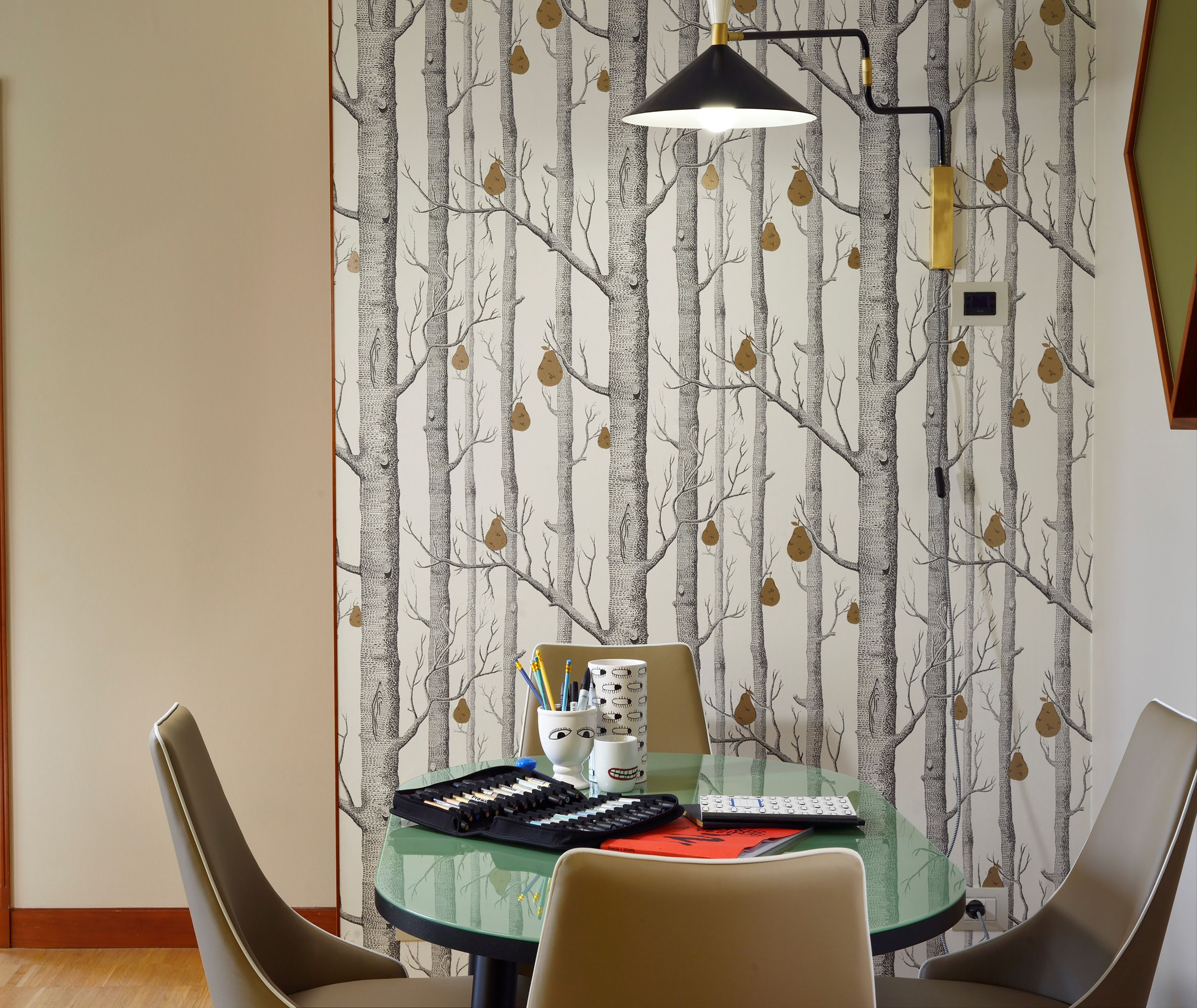 Un comedor con una mesa redonda de cristal, dos sillas, una lámpara y una pared decorada con un diseño de árbol.