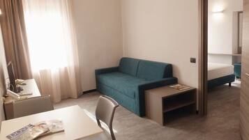 A compact hotel room with a single bed, a sofa, a small table, a chair, and a desk with a magazine.