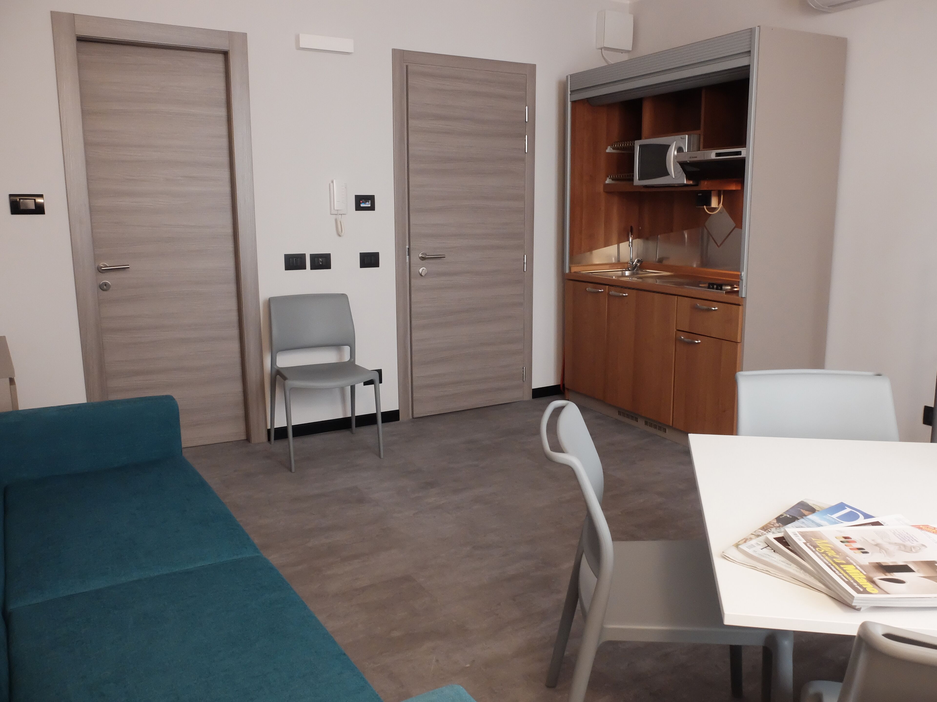 A room with a teal sofa, a dining table with magazines, a kitchen area with wooden cabinets and a microwave, and two doors.