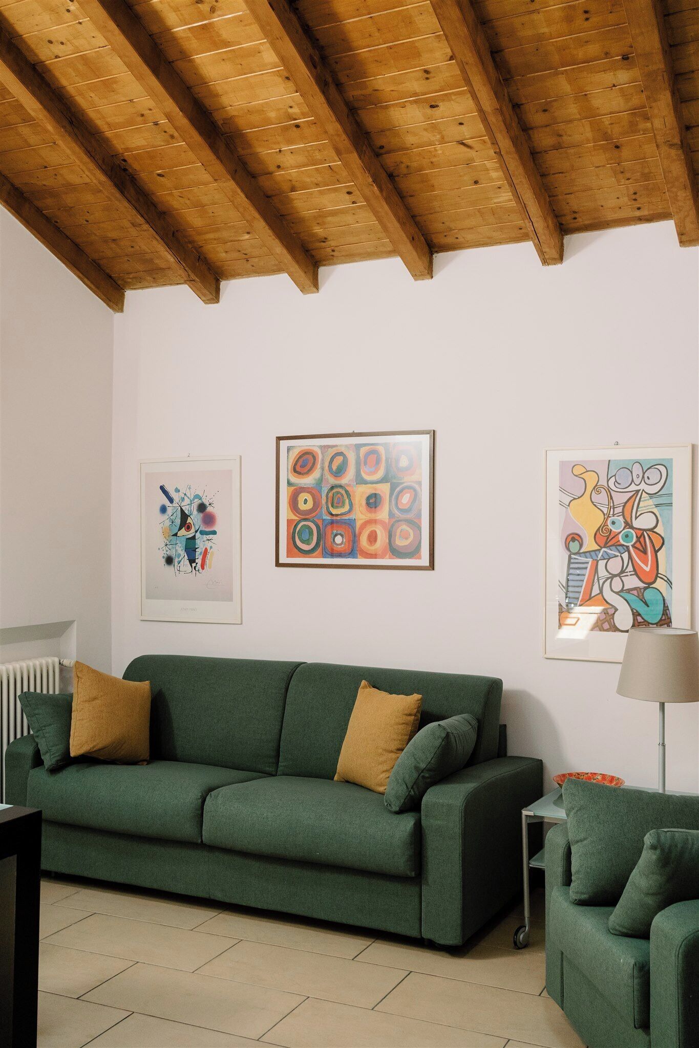Um sofá verde com duas almofadas, uma mesinha e um abajur em um quarto com teto de madeira e três obras de arte emolduradas na parede.