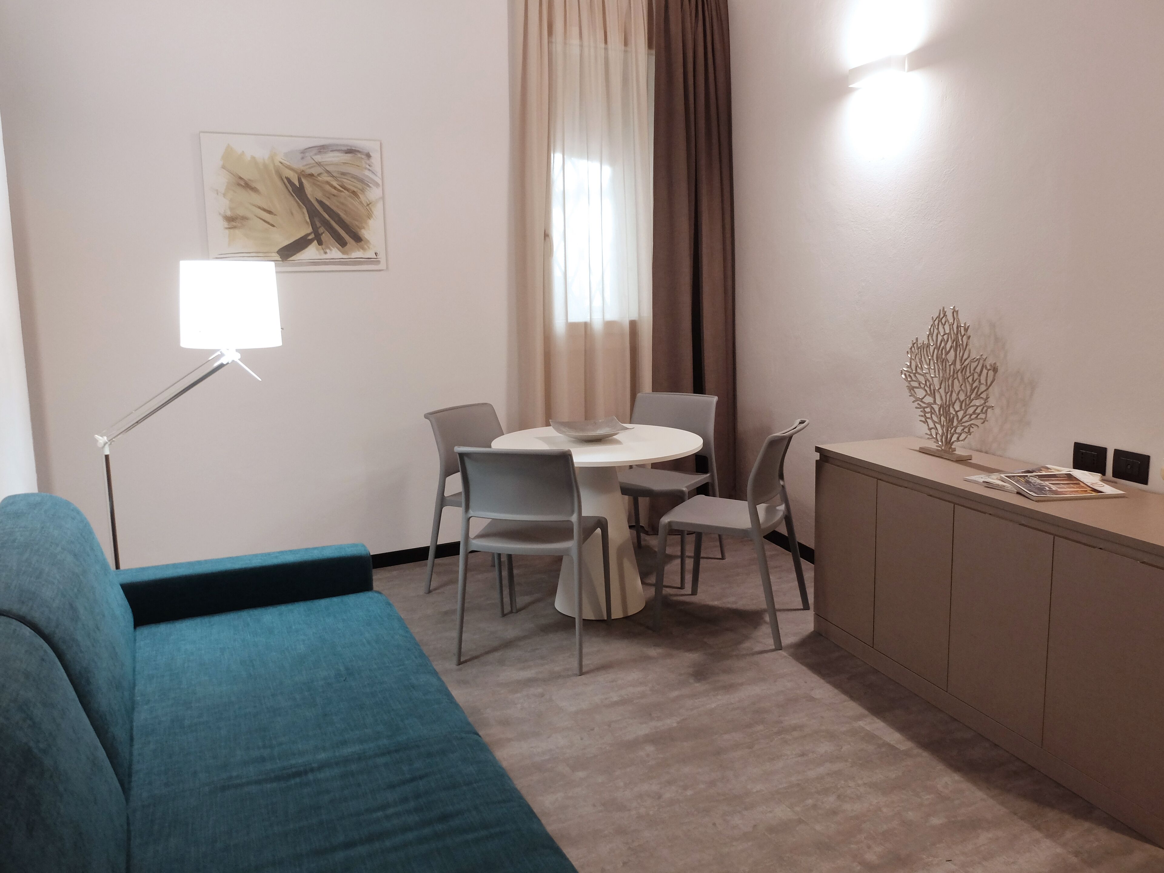 Una habitación de hotel moderna con una mesa redonda de comedor, sillas grises, un sofá azul, una lámpara de pie y una planta decorativa sobre un mueble.
