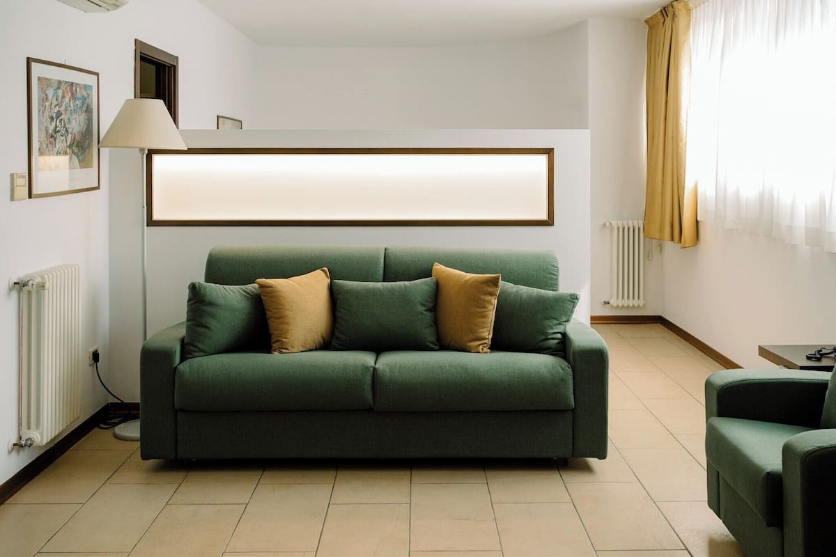 Un canapé vert avec deux coussins, un lampadaire, un tableau encadré au mur et une fenêtre avec des rideaux.