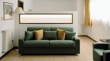 Un canapé vert avec deux coussins, un lampadaire, un tableau encadré au mur et une fenêtre avec des rideaux.