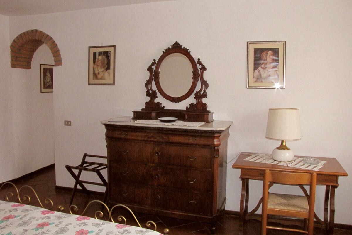 A bedroom with a wooden dresser, a mirror, a table with a lamp, a chair, and a bed with floral bedding.