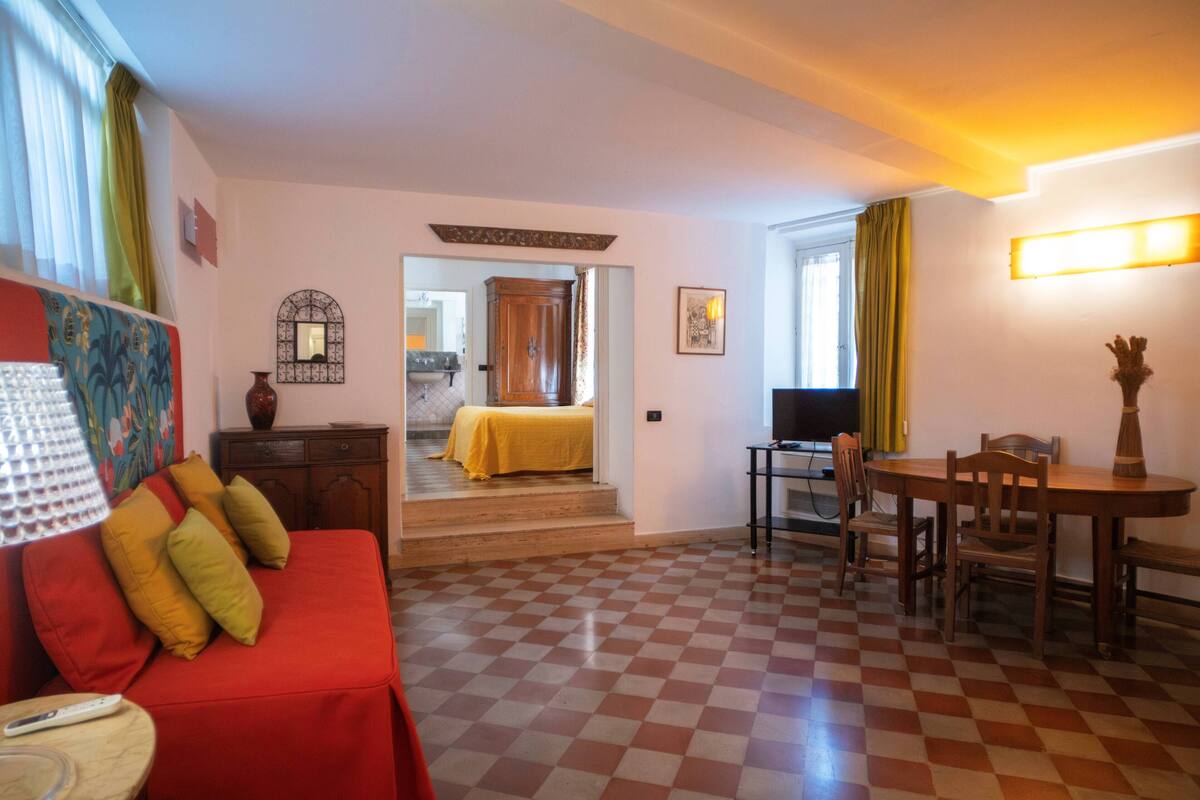 A hotel room with a checkered floor, a red sofa with pillows, a TV, a dining table with chairs, and a staircase leading to another room.
