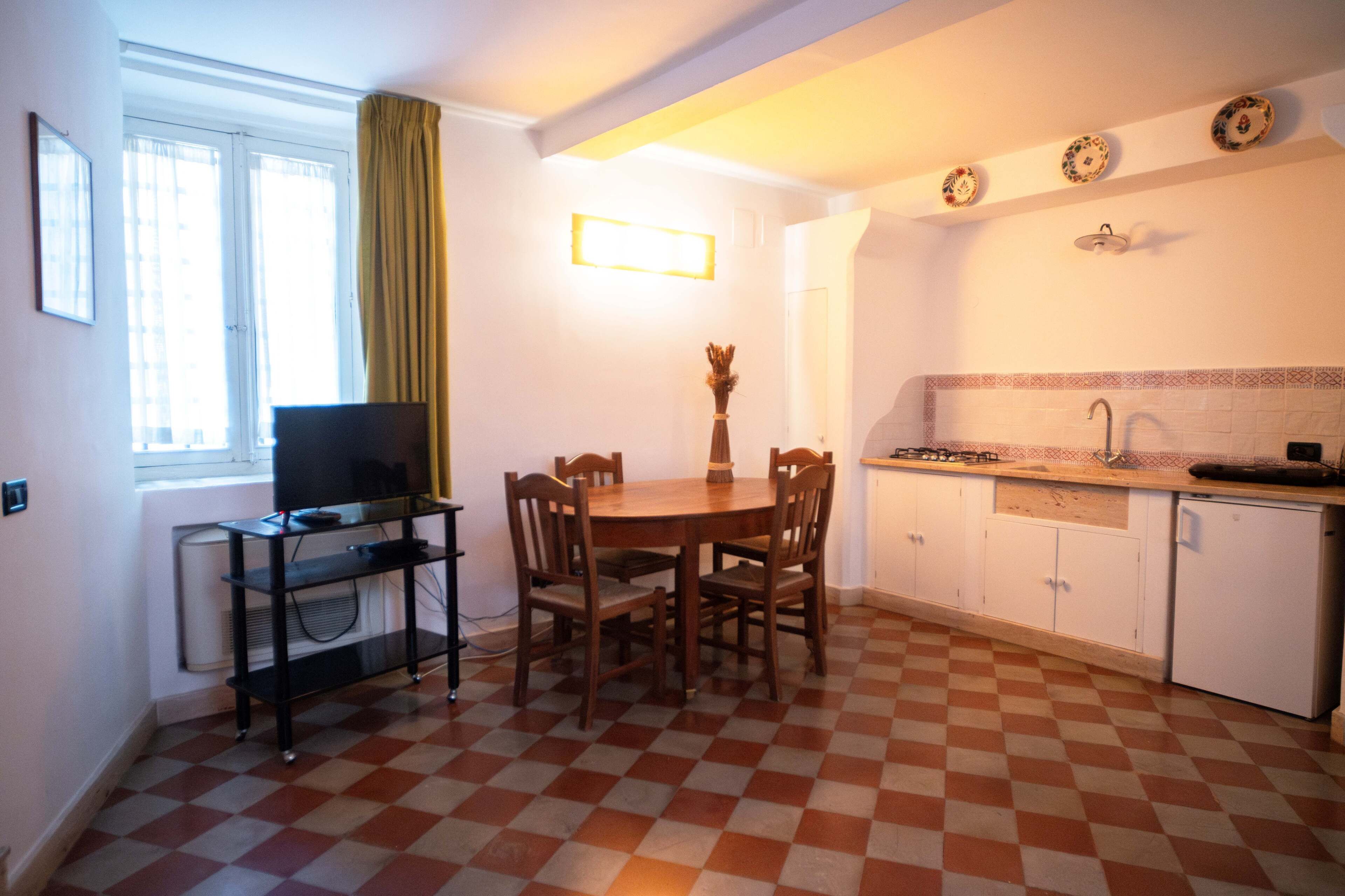A compact kitchen with a checkered floor, a dining table with chairs, a small refrigerator, and a window with curtains.