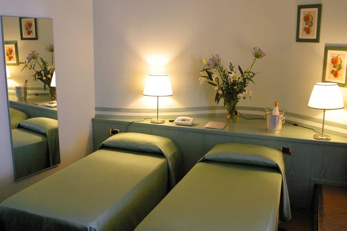 A hotel room with two single beds, a bedside table with a lamp, a mirror, and a vase with flowers.