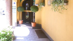 A narrow corridor with a yellow wall, two hanging lanterns, potted plants, and a door with a green sign. - Albergo della Corona (Binasco)