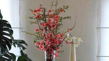 Ein Zimmer mit großem Fenster, einer Topfpflanze mit rosafarbenen Blüten und einer hohen weißen Vase.
