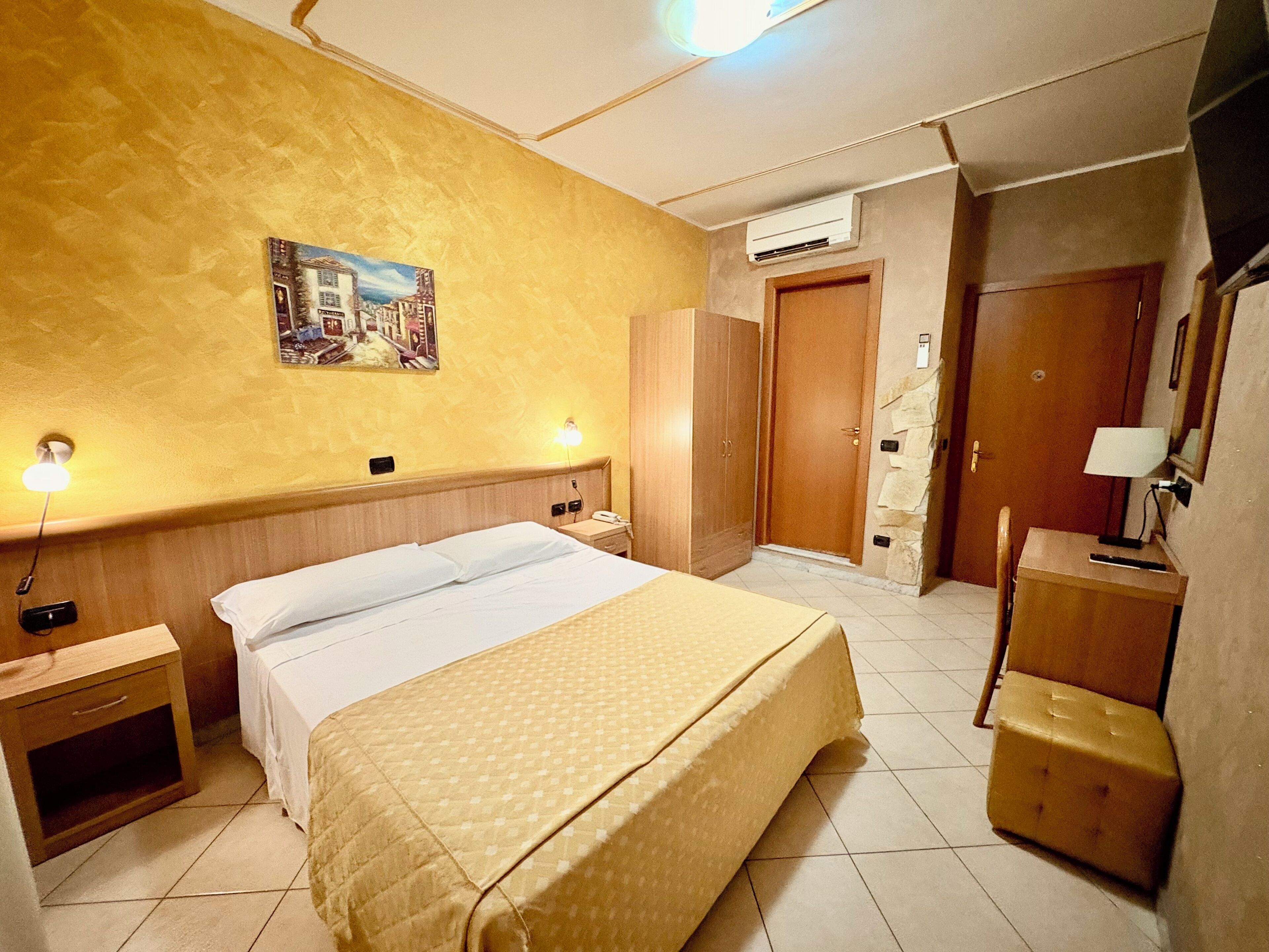 A hotel room with a bed, bedside tables, a wardrobe, a desk with a lamp, and an air conditioning unit.