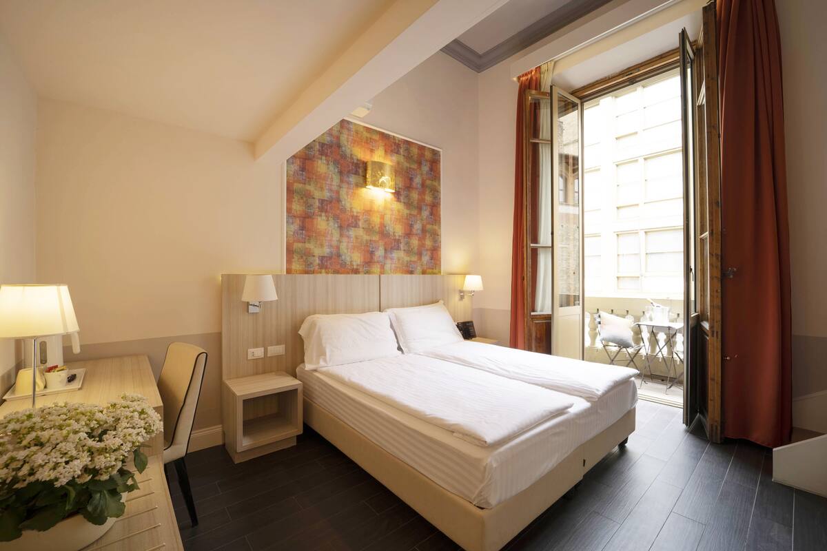 A hotel room with a large bed, bedside tables, a small desk, and a window with curtains.