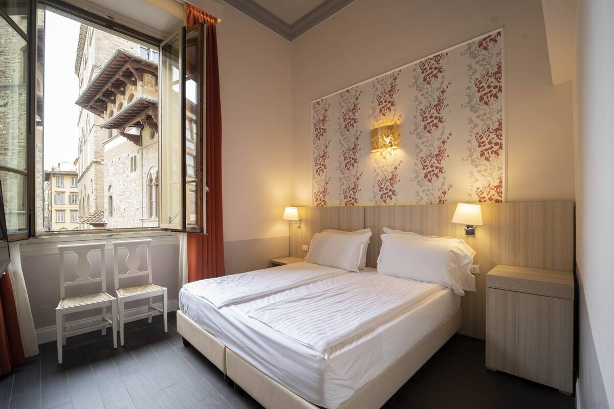 A hotel room with a large bed, two bedside tables with lamps, a window with curtains, and a view of buildings outside.
