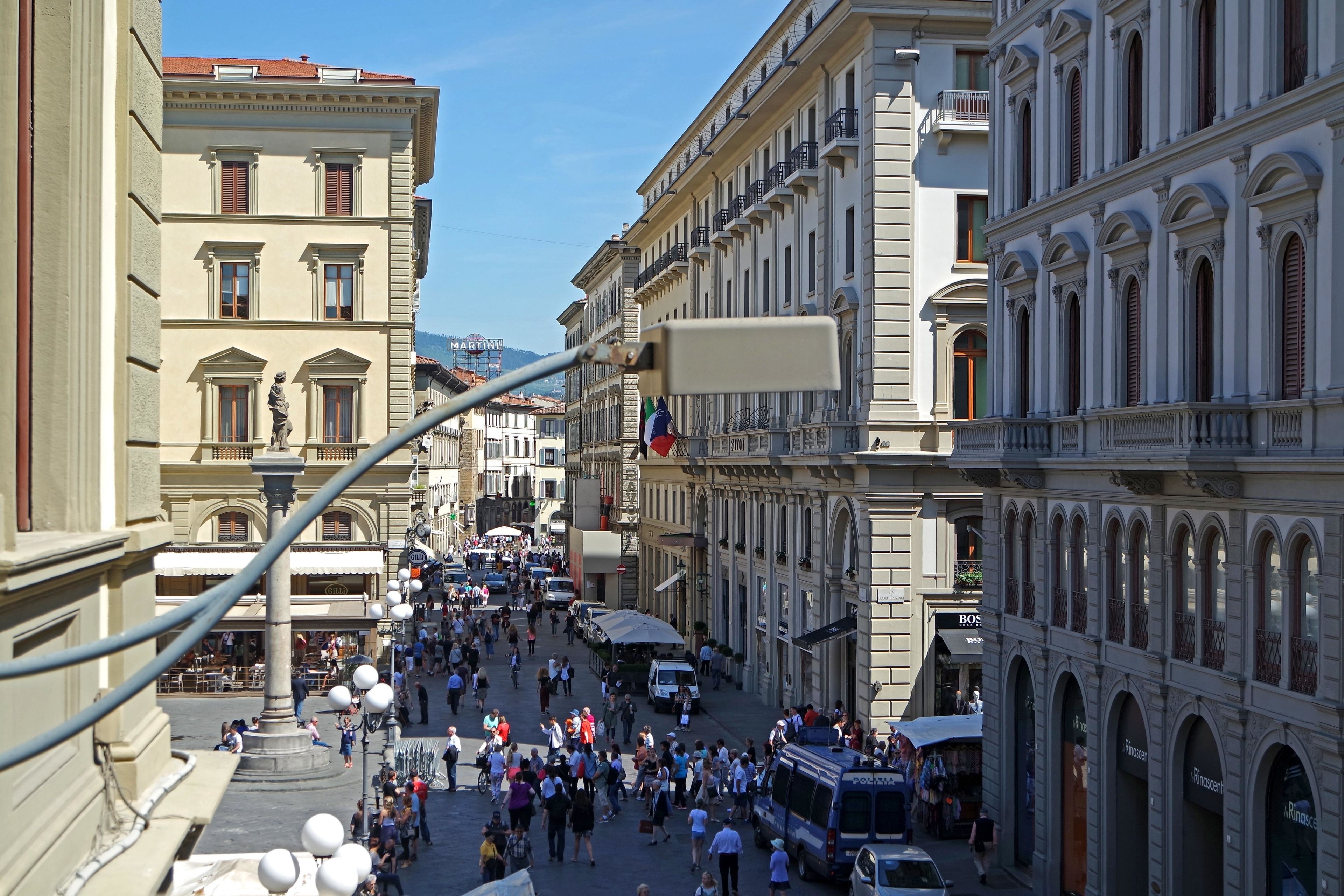 A busy urban street with historic buildings, outdoor seating, and a clear sky.
