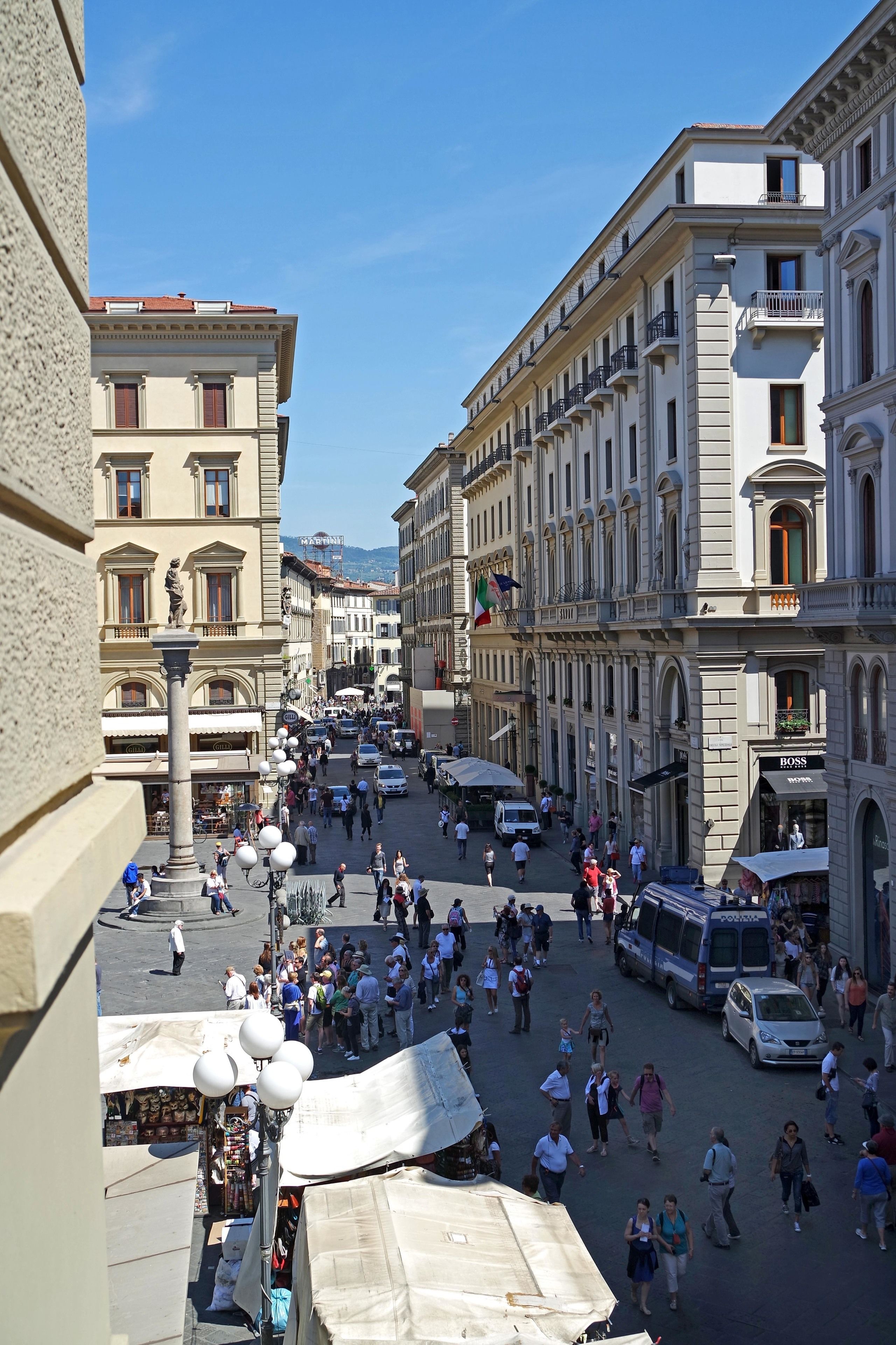 Una calle urbana concurrida con edificios históricos, una fuente central y una zona de mercado.