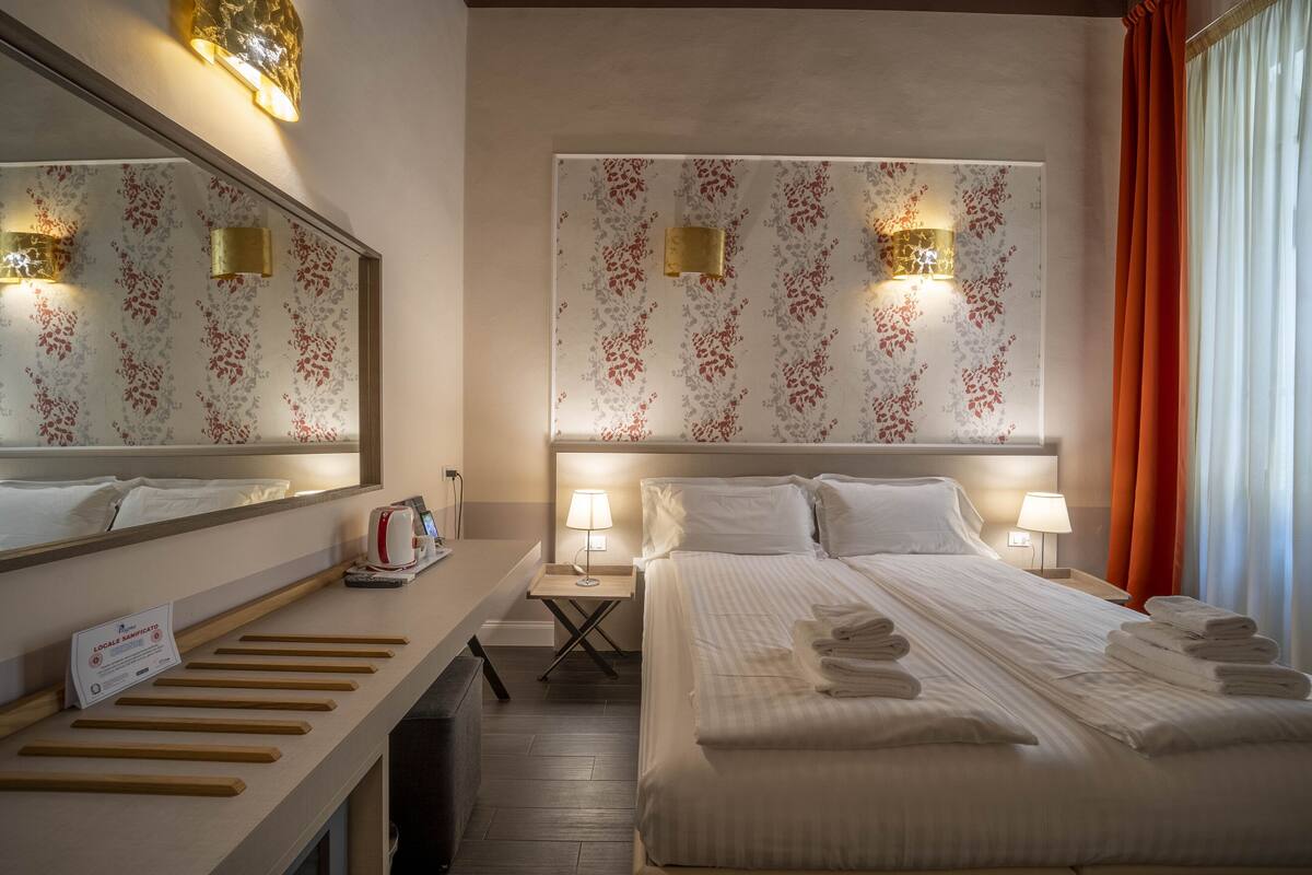A hotel room with a large bed, two bedside tables with lamps, a mirror, and a sign indicating local amenities.