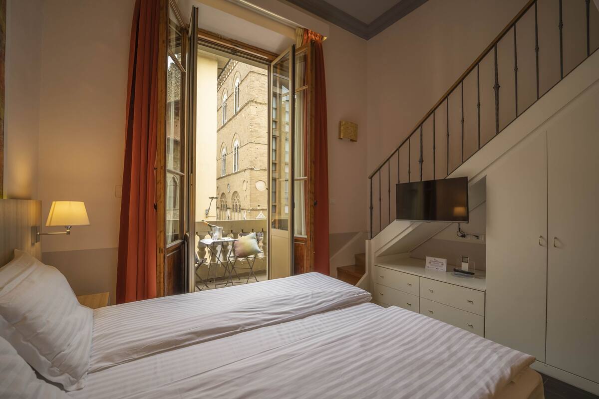 A hotel room with a bed, a desk with a lamp, a TV, and a view of a building through an open door.